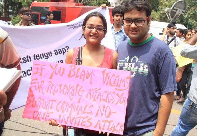 An Indian woman and man smile and hold a protest sign that says: You blame victims