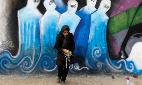 A young girl in black hijab looks down, while standing in front of a mural, with women in blue and white chadors