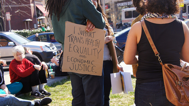 Two women stand at a protest, while other people sit on the grass in the background. They hold a sign that says: I support transgender equality and economic justice