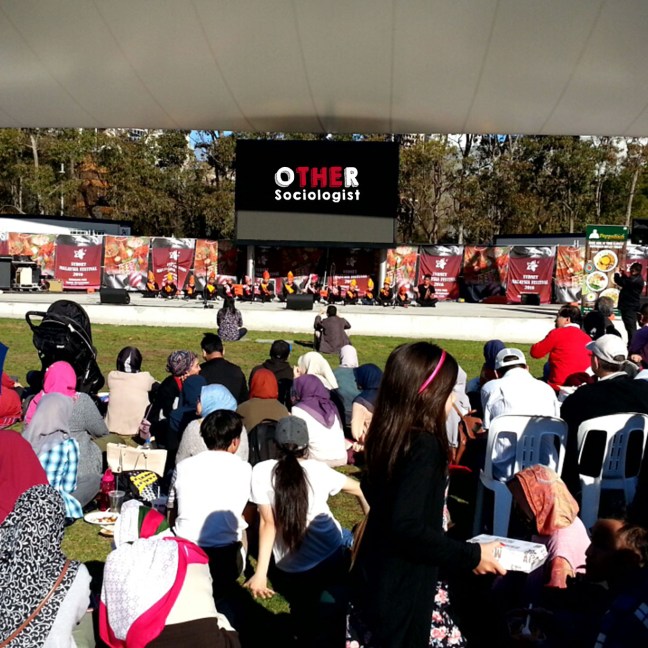 A community of migrants sit on a lawn, watching performers on a stage in the distance