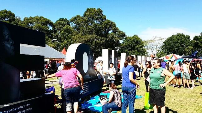 Women stand outdoors on a sunny day, in front of a life-sized sign that says Equality