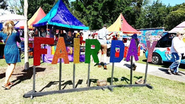 A fair in the background, with people walking, stalls, and a van. A sign says: Fair Day
