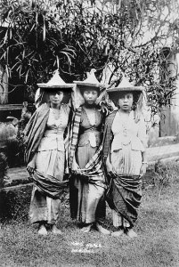 Three women stand together. They wear pointed hats, fitted vests, billowy pants and long fabric wrapped around their backs and scross their lower legs
