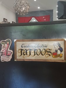 1950s tattoo artwork displayed at a black counter. Shows a cobra saying 'Cash only.' The larger sign shows a cartoon woman with a banner that reads, "Trixie," and the larger lettering: Custom Electric Tattoos. AtBroadway Tattoo, Sydney