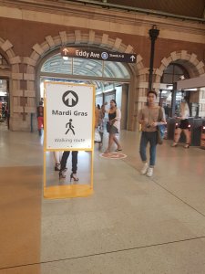 People walk in the background inside the grand Central Station. A sign shows an arrow and a person walking. It says: "Mardi gras walking route." In the background is the Eddy Ave exit sign