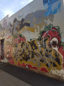 Street art. A large building has darwing of fierce tomatoes with bright white fangs, angry eyes and its vines look like large claws. On the botoom, we can see the artist's name, Ellice