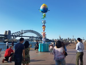 People stand in the foreground, waiting patiently and respectfully for a turn to take their photo with a tower of five cute, brightly-coloured monkeys standing on top of one another's heads. In the background is the iconic Sydney Harbour Bridge against a clear, blue-sky. The monkeys are juggling a gaint blue and yellow peach at the top of their primate tower