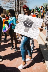 Young Black woman holds a sign with small writing and a large Black power sign. In the background are other protesters, one of them is wrapped in the Pride flag