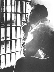 Martin Luther King Jr stares out of a jail cell. He is bathed in light. He has his elbows resting on his knees and his hands on his chin. He looks up and is pensive