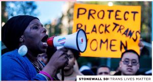 A Black woman yells into a megaphone. In the background is a yellow sign with black writing that partially reads: Protest Black