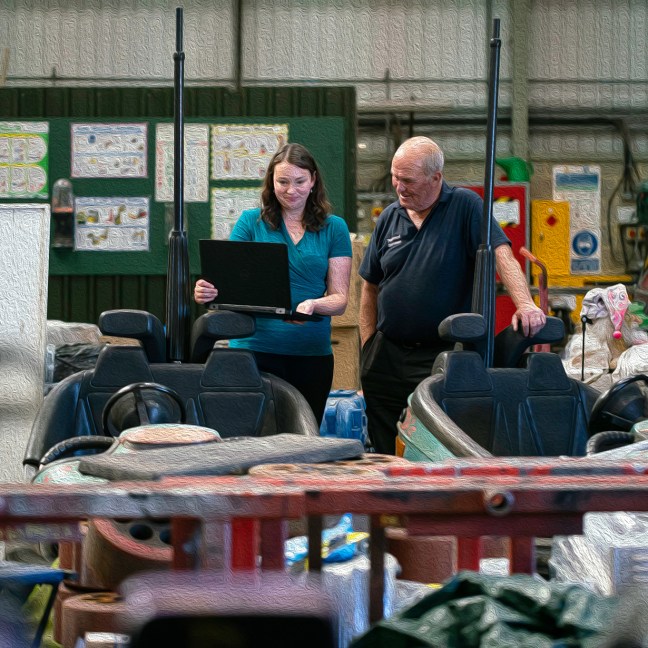 A young white woman stands next to an older white man in an industrial workshop. They are both smiling looking at her laptop