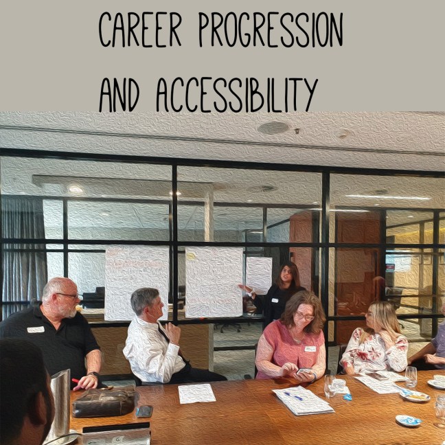 Three men and one woman sit at table. They watch another woman who is pointing to the group notes on a wall