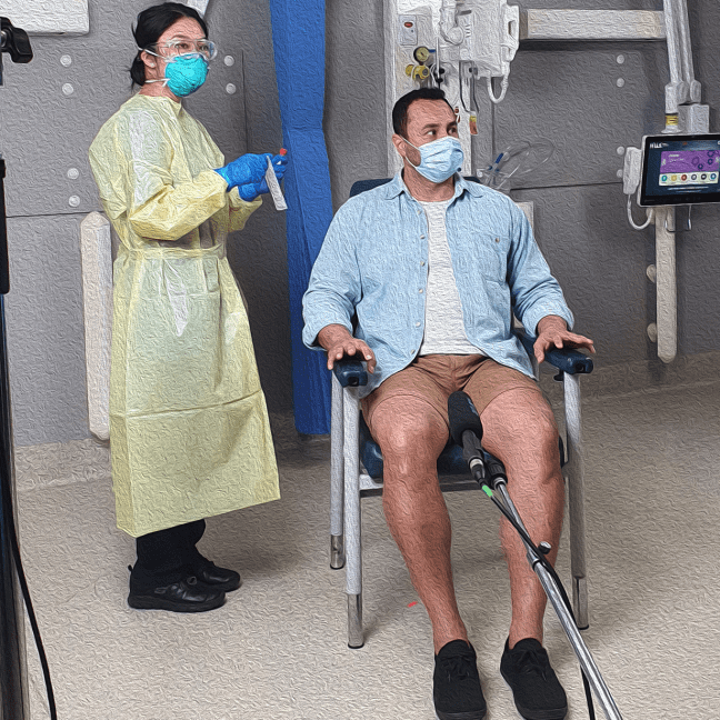 An Asian woman clinician stands, holding a COVID PCR test. She wears a surgical mask and gown. A Pacific Islander man sits in a chair wearing a mask. The both look to the side as if listening to instructions