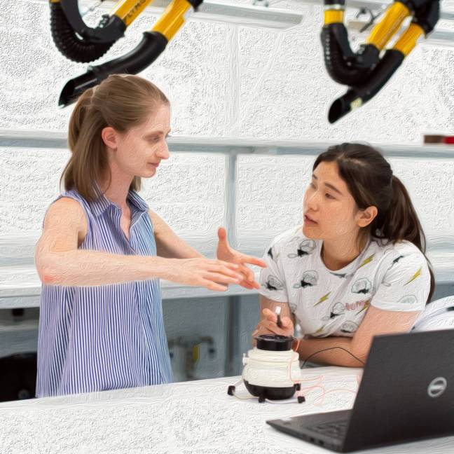 A white woman is gesturing with her hands while an Asian woman listens. They are in an engineering lab with a white machine and laptop in front of them
