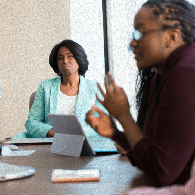 A Black woman in a jacket watches another Black woman talking
