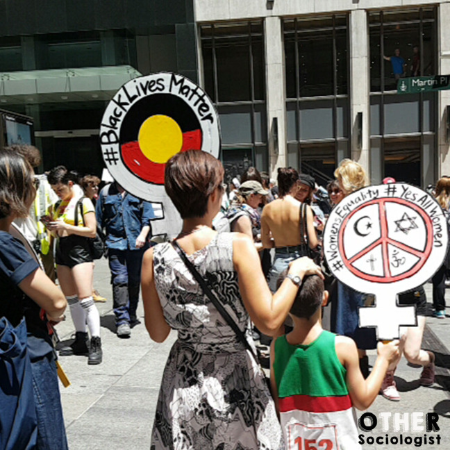 crowd at the Sydney Women’s March, with a woman holding a “Black lives matter” sign bearing the Aboriginal flag, with her hand on the head a young boy who holds a sign made from the symbol for “woman,” bearing religious symbols for Islam, Judaism, Christianity and Hinduism, with the words, #WomensEquality and #YesAllWomen