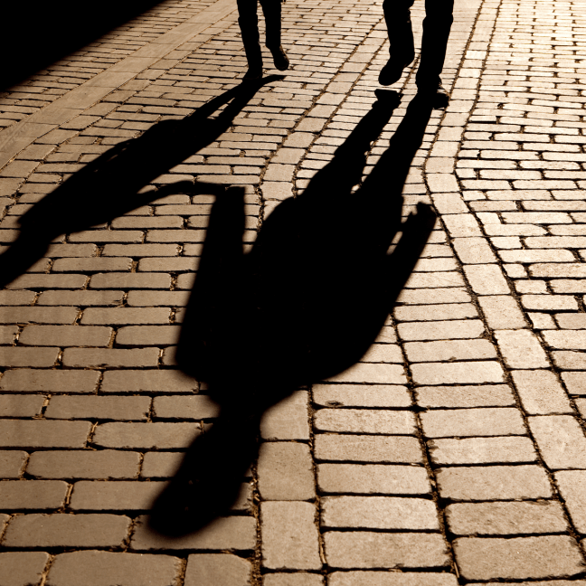 View of bricked ground with two shadows looming, with two sets of feet in the background