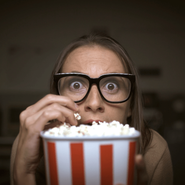 Young white woman wearing glasses hides behind a bucket of popcorn