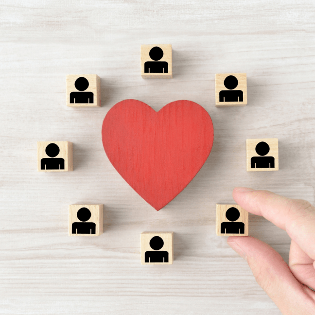 A man's fingers holds a block with a person icon. The blocks are arranged around a wooden love heart