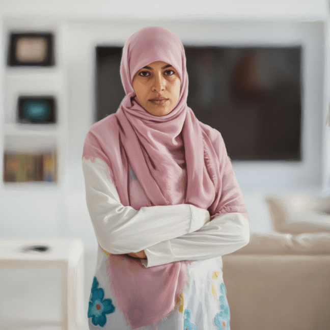 A Middle Eastern woman wearing a pink hijab stands in a room, with her arms crossed