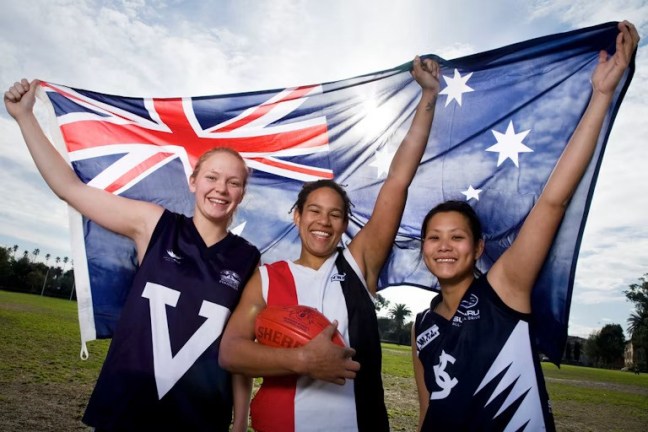 Three girls in footy uniform hold up the Australian flag in triumph. One is white, the other Indigenous, and the third is Asian