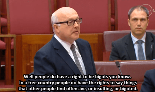 George Brandis arguing in Parliament. He is a white, balding man who wears glasses and a blue suit. He says: Well people do have a right ot be bigots you know. In a free country people do have the rights to say things that people find offensive, or insulted, or bigoted