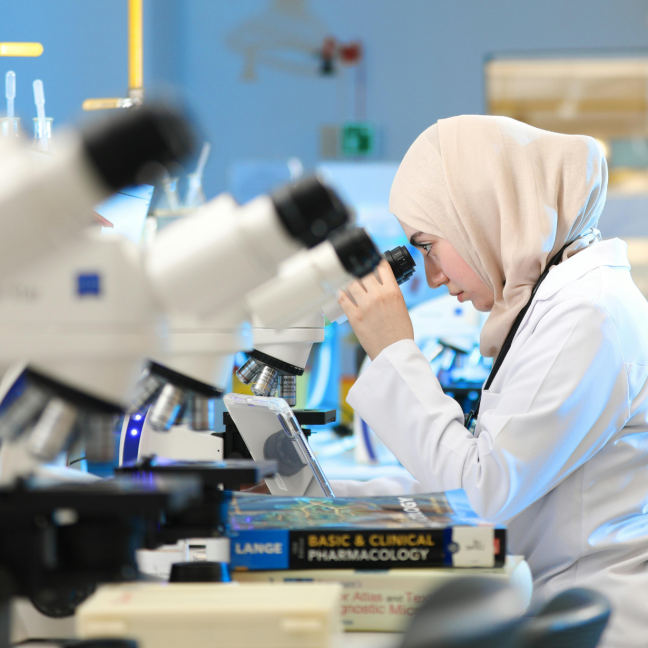 An Asian Muslim woman in hijab and a lab coat looks into a microscope