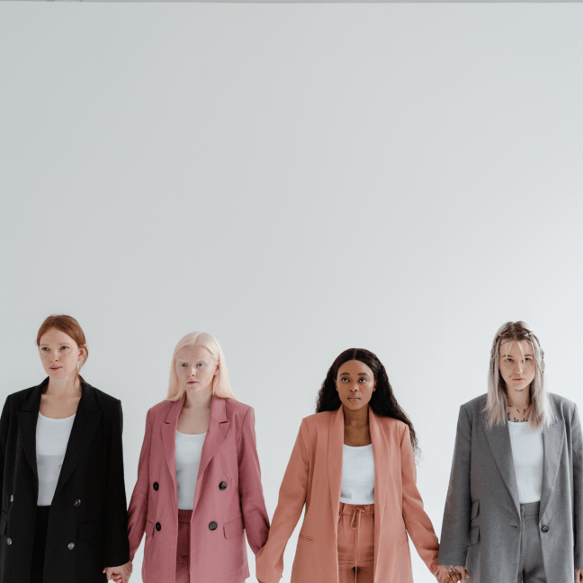 Four young women wear suits and hold hands, one of them is very fair, one woman is Black, and the other two are white women, one of them covered in neck and chest tattoos