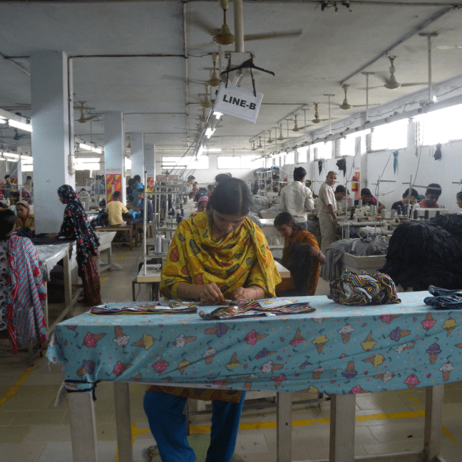 A Bangladeshi woman sews clothing on a long narrow table, in a factory filled with many workers standing and sitting as they work