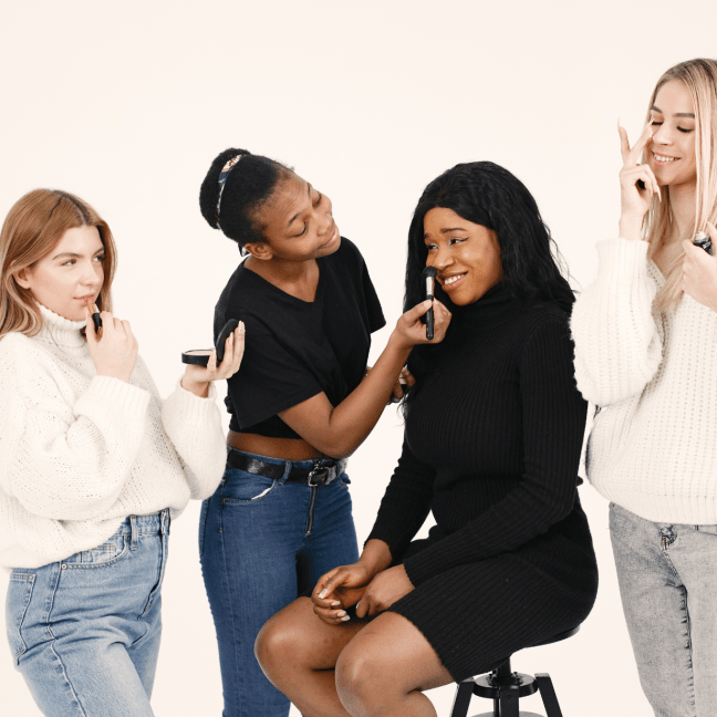 A young Black woman does the make up for another Black woman who looks unsure, while two while women sput on make up on either side of them