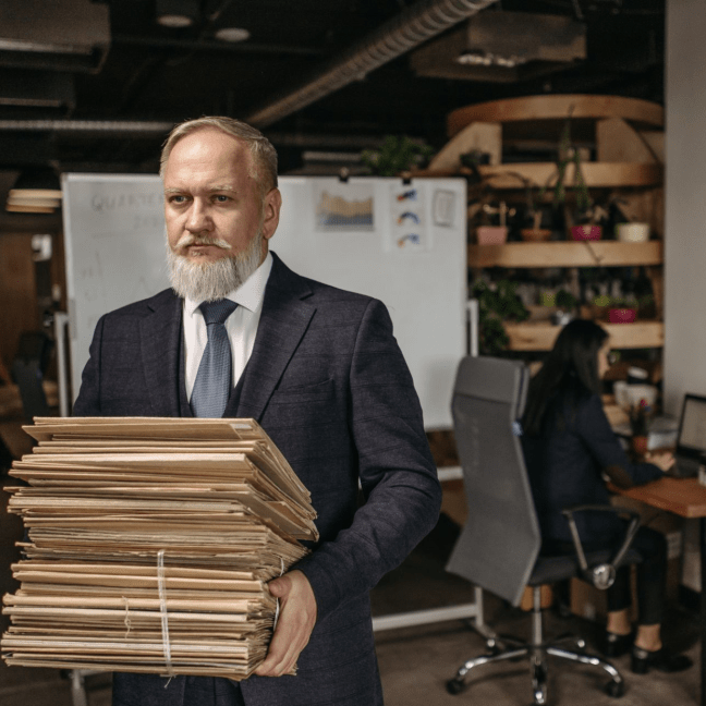 An older white man with a beard and moustache carries a large pile of files, while a woman works on a computer in the background