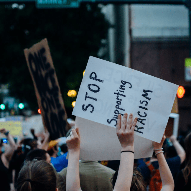 A crowd of protesters hold sign. White women's hands hold up a sign that says: Stop supporting racism