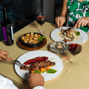 Two people seen from neck down as they eat a meal with a bottle of red wine and glasses on the table