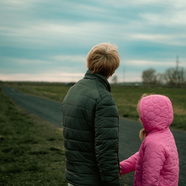 Parent and daughter seen from behind, standing on a rural path on a cloudy day