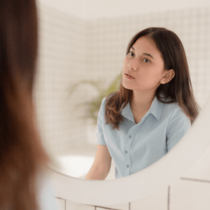 An Asian woman stares into a bathroom mirror with a haunted expression