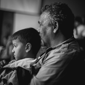 Black and white photo of a Middle Eastern grandfather kissing the back of his grandson's head, who is sitting on his lap and looking ahead with concentration