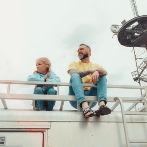 A father and daughter sit together on a rooftop, squinting as they look to the side