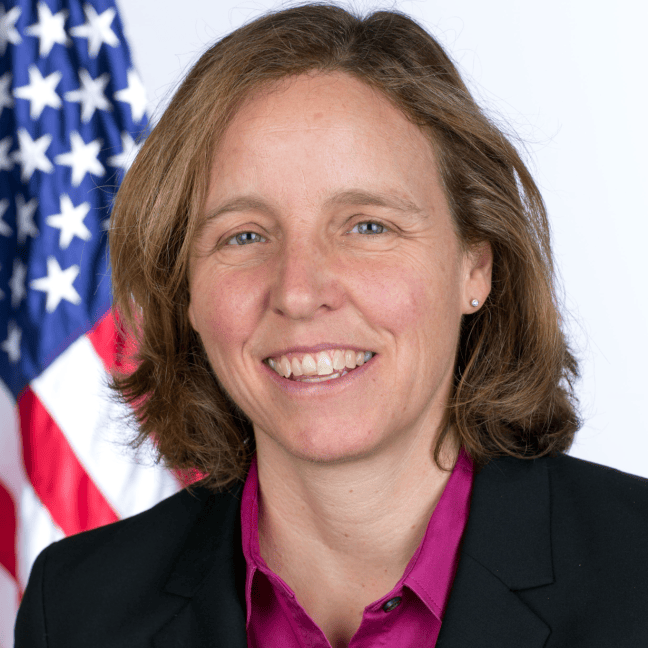 Megan Smith is a white woman with short dark blonde hair and blue eyes, who wears a black jacket and fuschia shirt. She smiles. Behind her is the USA flag.